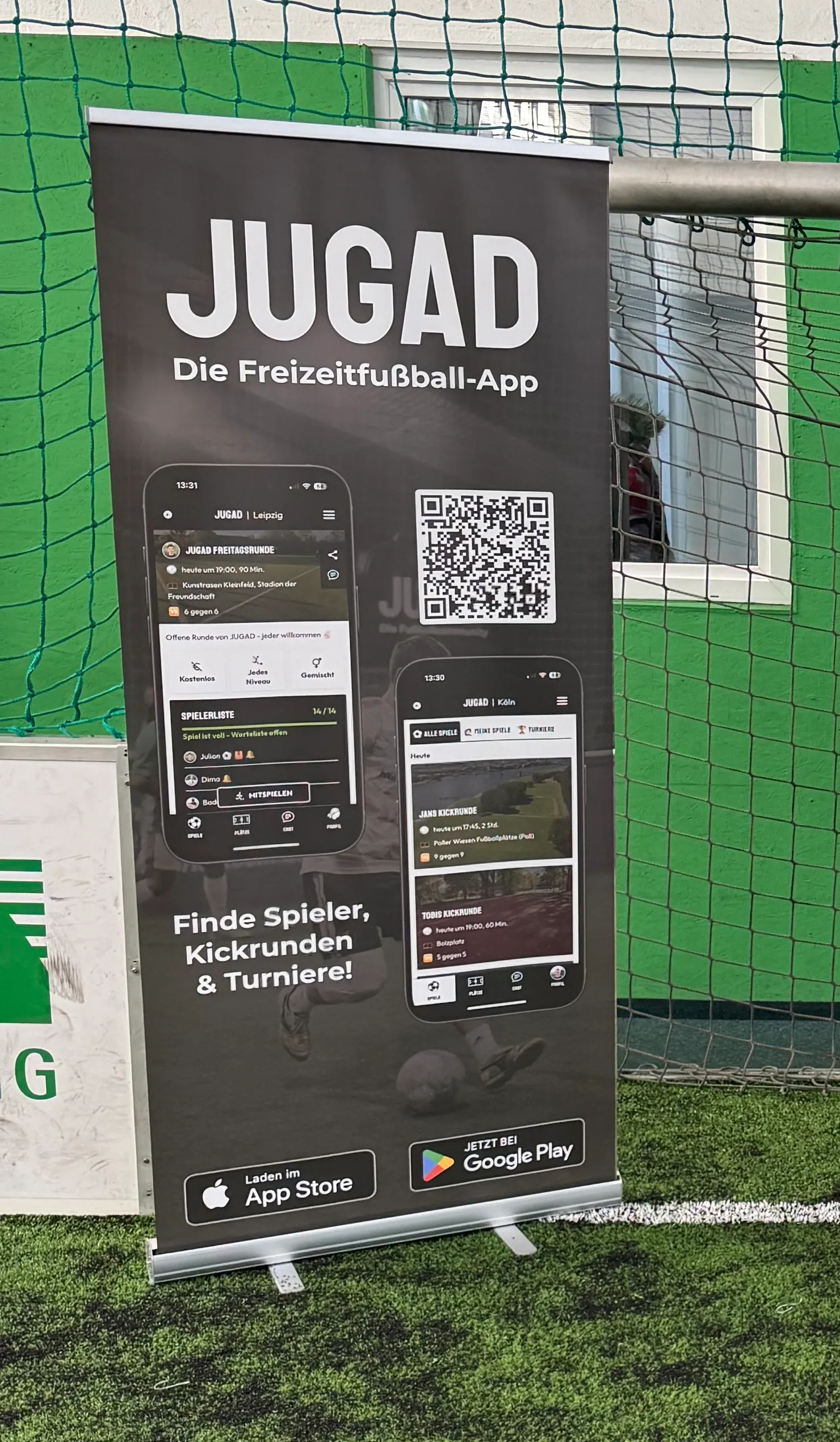 JUGAD-Aufsteller „Die Freizeitfußball-App“ mit App-Screenshots, QR-Code und Store-Buttons, aufgestellt auf Kunstrasen vor einem Fußballtor